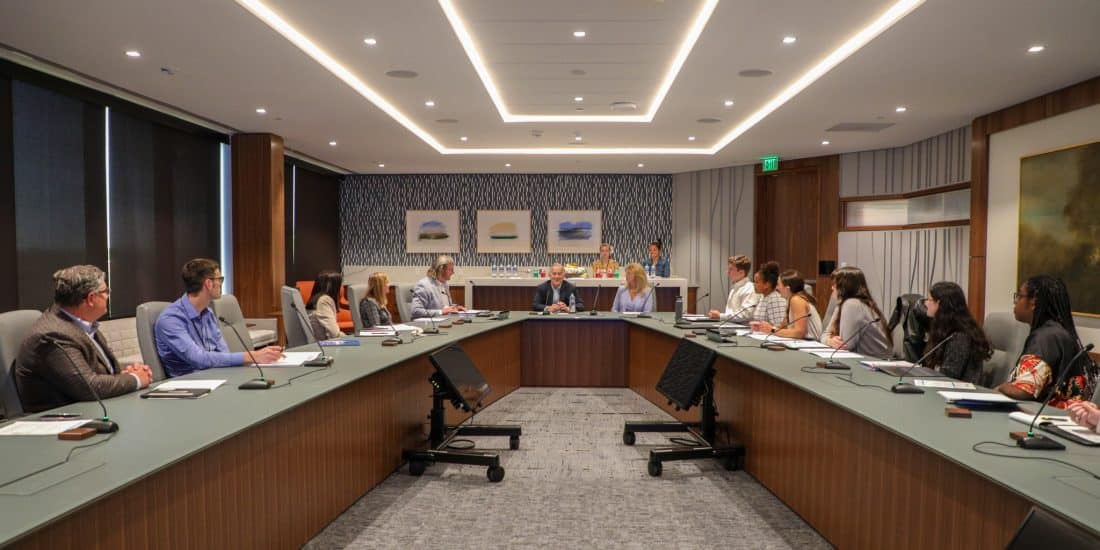 A group of students sitting around a long board room table with two executives at the head of the table