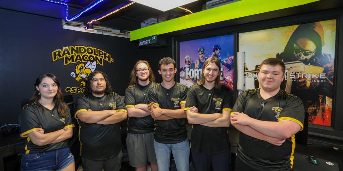 Group of six individuals in esports jerseys, posing with arms crossed in a gaming lounge with neon lighting and game posters in the background.